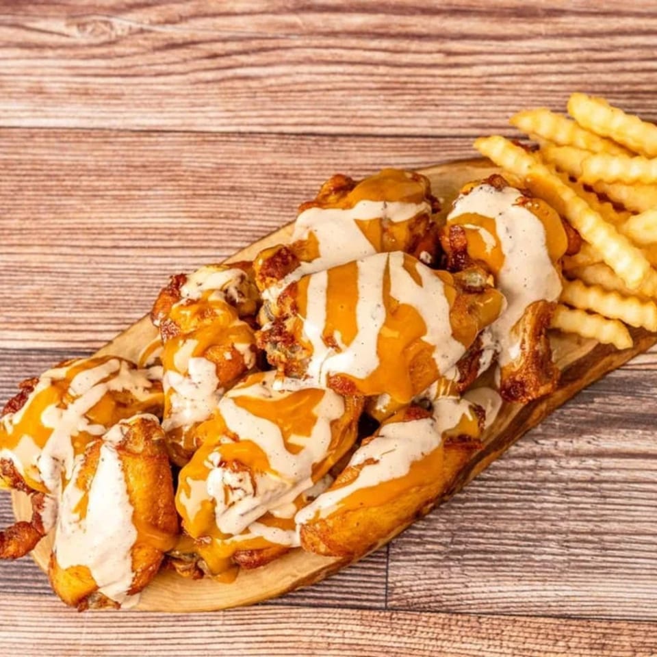 Chicken wings drizzled with orange and white sauces, served with crinkle-cut fries on a wooden board.